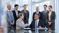 
  Several business people standing around a table watching two people sitting down shaking hands
