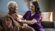 
  Healthcare provider sitting on a couch with a patient in their home checking their vitals. 
