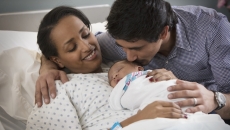 
  A person laying in a hospital bed with a newborn on their lap and another person standing beside them
