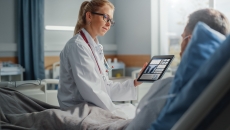
  Healthcare provider sitting on a patient's hospital bed while holding a tablet showing diagnostic images of lungs
