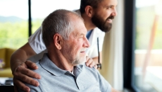 One person sitting in a chair while another person stands beside them as both look out of a window