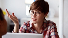 
  A person with a red plaid shirt on with short hair wearing glass, sitting at a computer with their hand up while talking
