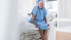 
  Patient sitting on the side of a bed in a hospital room wearing a hospital gown 
