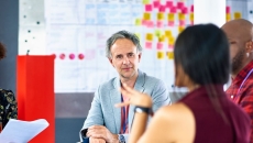
  People sitting around a table in front of a whiteboard talking to each other 

