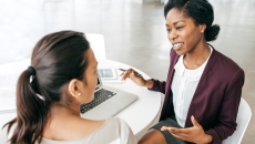 Two people in business attire chat at a table holding a laptop