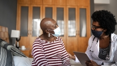 Two individuals sitting in a home looking at each other while wearing face masks