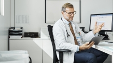 Healthcare provider sitting in a chair by a desk with a computer on it while talking to someone on a tablet