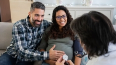 Two people sitting on a couch with one rubbing the other's stomach while both speak to a healthcare provider