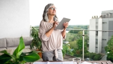 Person standing on a balcony holding a tablet while looking up at the sky with a city behind them