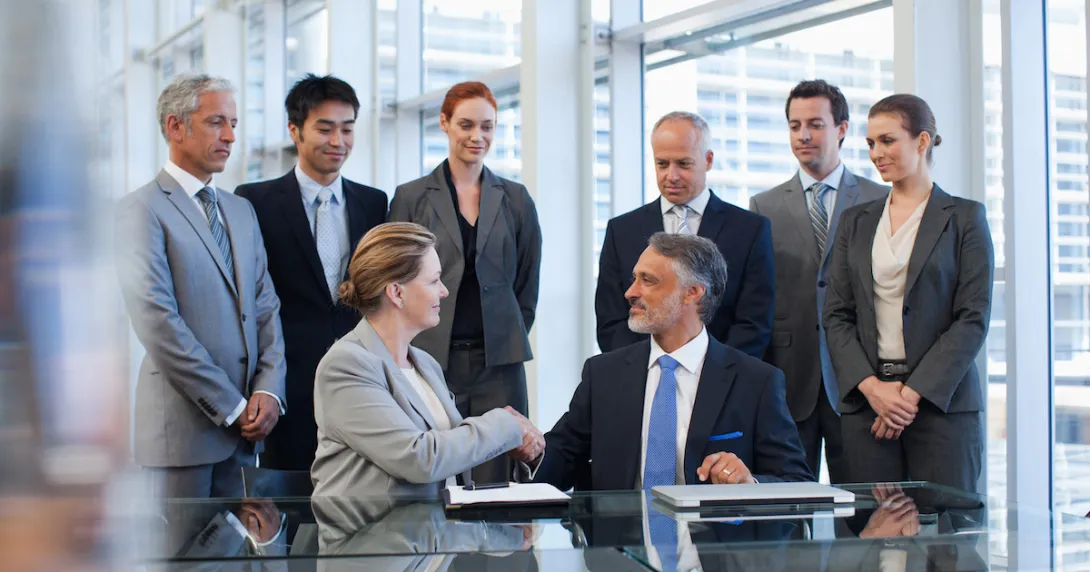 Several business people standing around a table watching two people sitting down shaking hands
Several business people standing around a table watching two people sitting down shaking hands