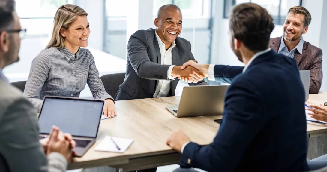 Five businesspersons sitting around a table with two of them shaking hands Five businesspersons sitting around a table with two of them shaking hands