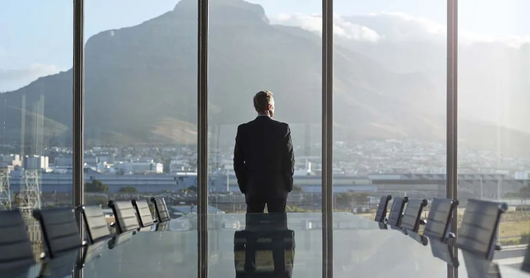 Person standing by a large window with their back turned looking at a large mountain
Person standing by a large window with their back turned looking at a large mountain