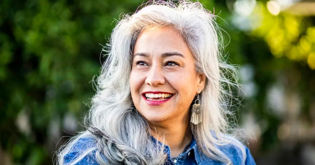 Person with long gray hair smiling while standing in front of trees Person with long gray hair smiling while standing in front of trees