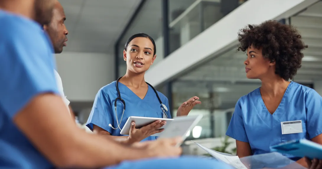 Four healthcare professionals wearing blue scrubs and sitting in a circle with one holding a tablet
Four healthcare professionals wearing blue scrubs and sitting in a circle with one holding a tablet