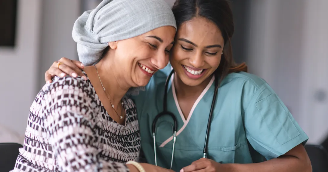 Two people sitting next to each other, one wearing a scarf on their head and the other in scrubs Two people sitting next to each other, one wearing a scarf on their head and the other in scrubs