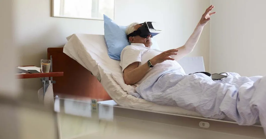 Person in a clinical setting lying in bed with their hand raised while wearing a virtual reality headset
Person in a clinical setting lying in bed with their hand raised while wearing a virtual reality headset