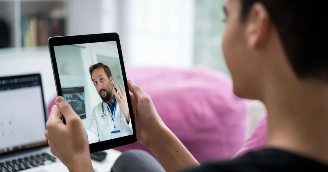 Person sitting in a room while holding a tablet that has a healthcare provider on the screen Person sitting in a room while holding a tablet that has a healthcare provider on the screen
