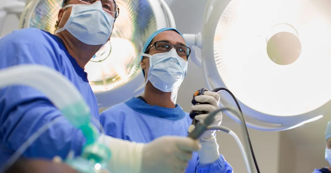Two surgeons looking down at a camera with scrubs on while holding surgical tools
Two surgeons looking down at a camera with scrubs on while holding surgical tools