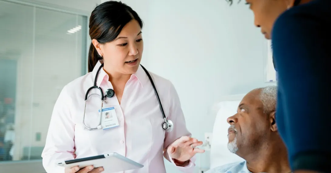 Doctor holding a tablet and standing by a patient and another individual Doctor holding a tablet and standing by a patient and another individual