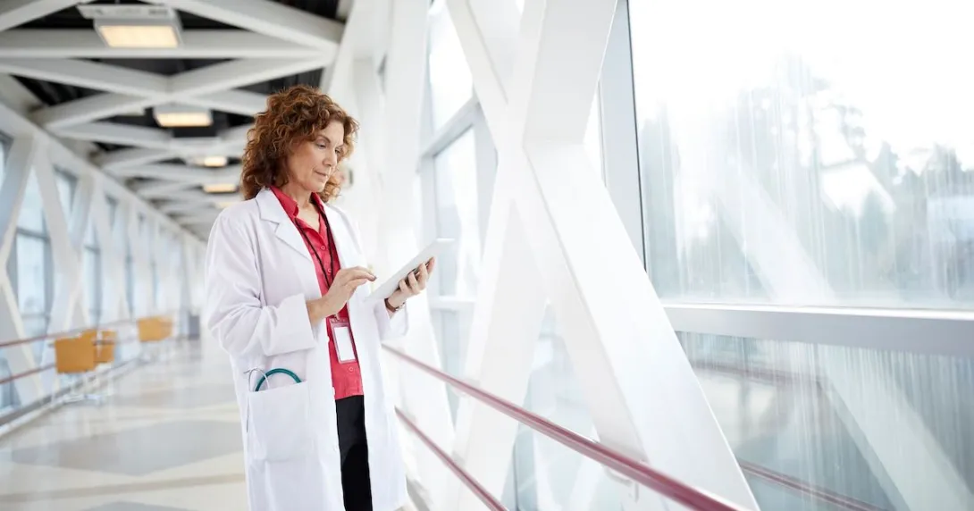 Healthcare provider holding a tablet while wearing a lab coat and standing in a hallway by a large window Healthcare provider holding a tablet while wearing a lab coat and standing in a hallway by a large window
