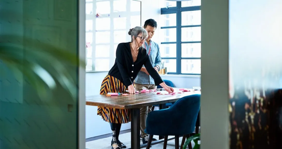 Two people standing in a room looking at something on a desk while standing above it
Two people standing in a room looking at something on a desk while standing above it