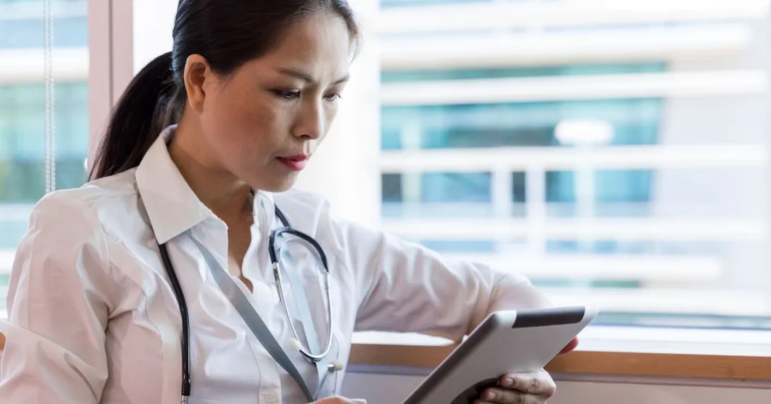 Person sitting by a window wearing a stethoscope and looking at a tablet Person sitting by a window wearing a stethoscope and looking at a tablet