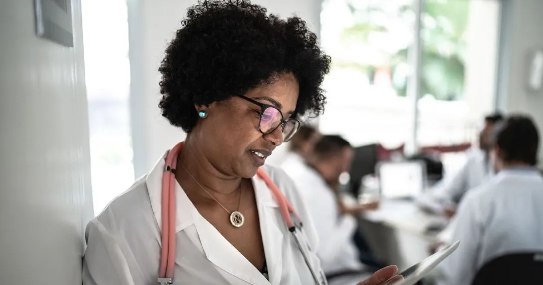 Healthcare provider standing against a wall and looking at something in their hand Healthcare provider standing against a wall and looking at something in their hand