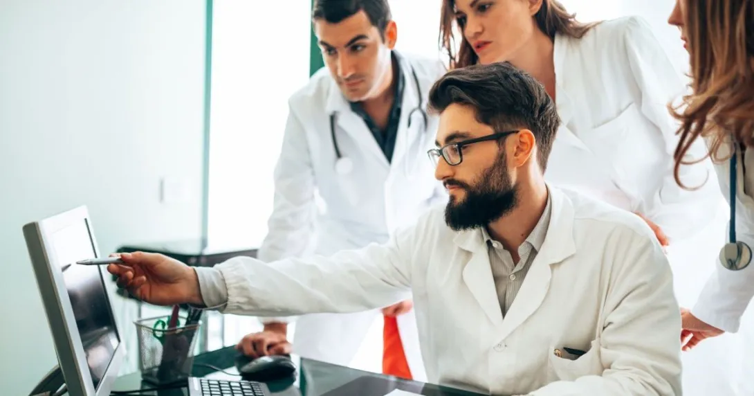 Three people wearing lab coats with one seated and pointing at screen Three people wearing lab coats with one seated and pointing at screen