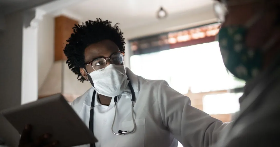 Healthcare provider wearing a mask, a lab coat and a stethoscope around their neck talking to a patient in their home
Healthcare provider wearing a mask, a lab coat and a stethoscope around their neck talking to a patient in their home