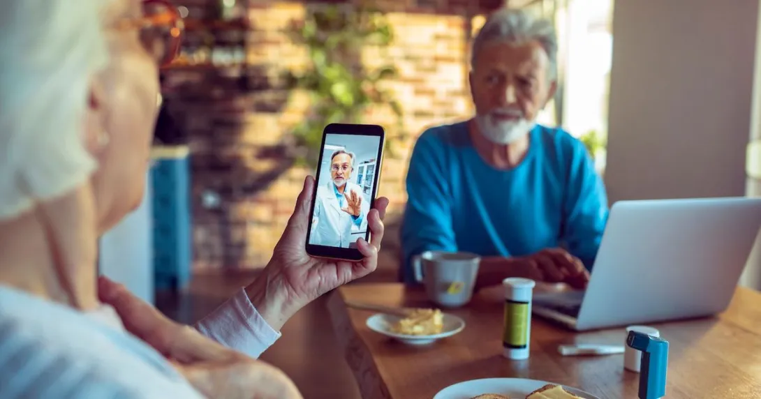 Two people sitting at a table with a computer on it and one of them is holding a phone
Two people sitting at a table with a computer on it and one of them is holding a phone