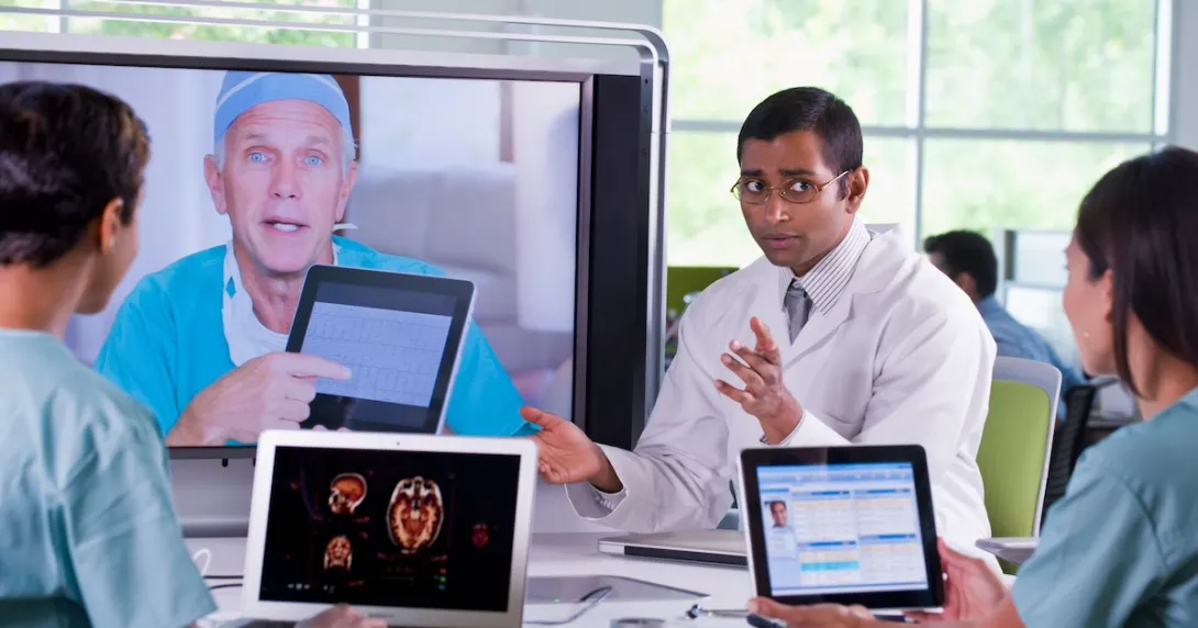 Healthcare team sitting around a table with laptops on it while talking to another healthcare provider on a TV screen Healthcare team sitting around a table with laptops on it while talking to another healthcare provider on a TV screen