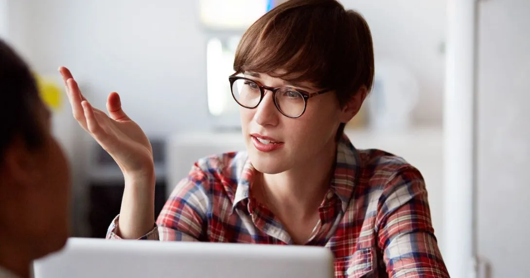 A person with a red plaid shirt on with short hair wearing glass, sitting at a computer with their hand up while talking
A person with a red plaid shirt on with short hair wearing glass, sitting at a computer with their hand up while talking