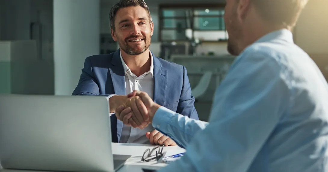 Two people sitting across from each other at a desk shaking hands Two people sitting across from each other at a desk shaking hands