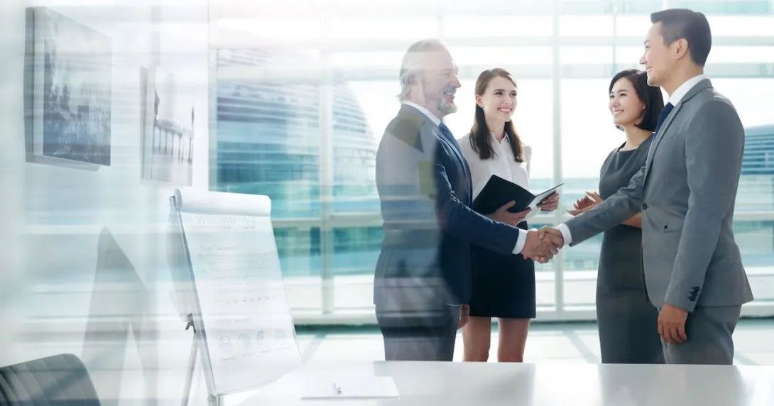 Four people standing in front of a large window with two of them shaking hands Four people standing in front of a large window with two of them shaking hands