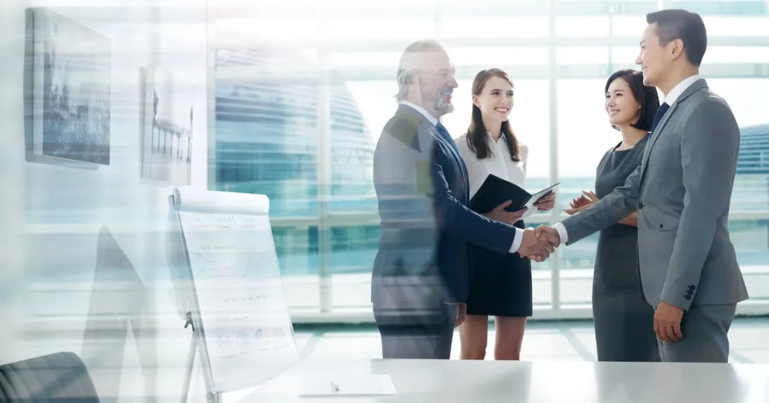 Four people standing in a room with two of them shaking hands Four people standing in a room with two of them shaking hands