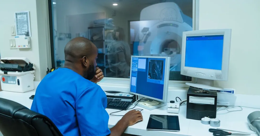 Healthcare provider sitting a room with a computer while there is a window for them to see a diagnostic imaging tool Healthcare provider sitting a room with a computer while there is a window for them to see a diagnostic imaging tool