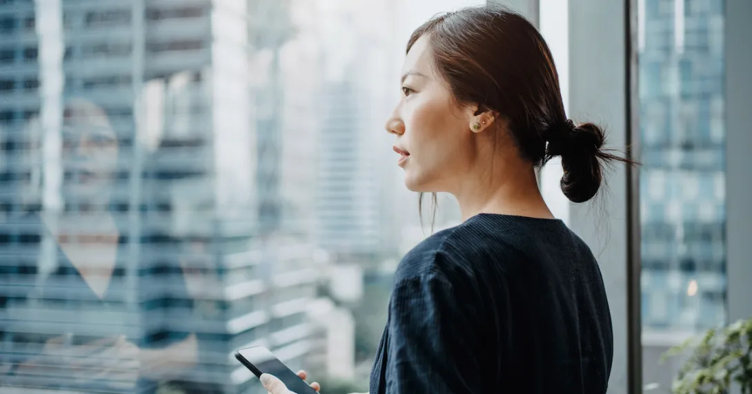 Profile shot of a woman holding smartphone and looking out an office window