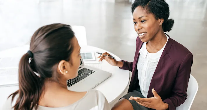 Two people in business attire chat at a table holding a laptop