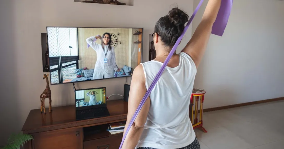 A woman watching and copying exercises with a resistance band in her living room, guided by a physical therapist online A woman watching and copying exercises with a resistance band in her living room, guided by a physical therapist online