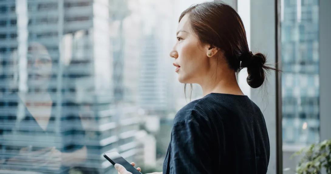 A woman holding a smartphone looking out a window. A woman holding a smartphone looking out a window.