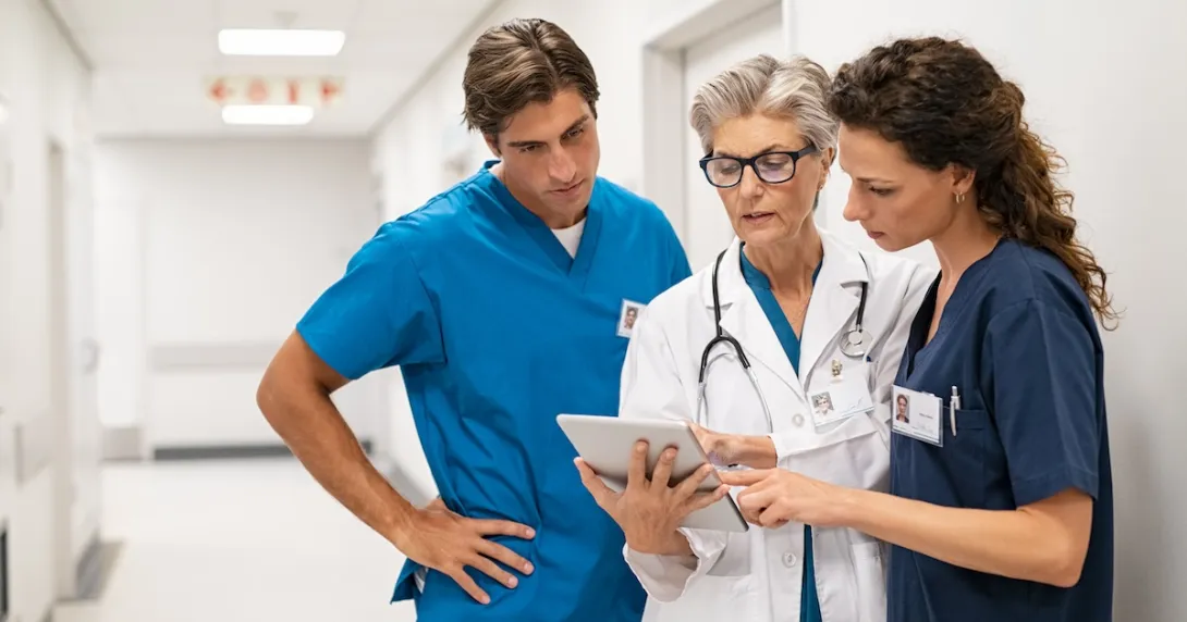 Three healthcare providers standing in the hallway of a clinical building looking at a tablet together