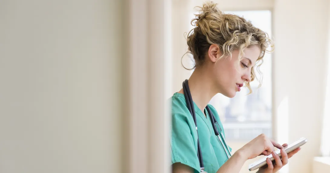 Healthcare provider standing in a hallway wearing green scrubs and a stethoscope around their neck while looking at a tablet