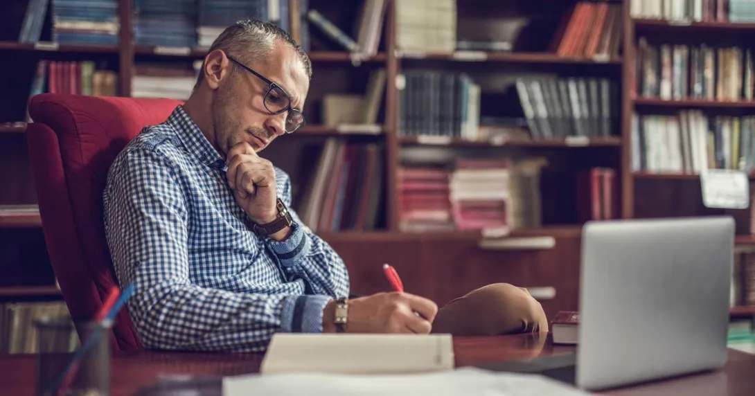 Person sitting at a desk writing something down on a piece of paper with a bookshelf behind them