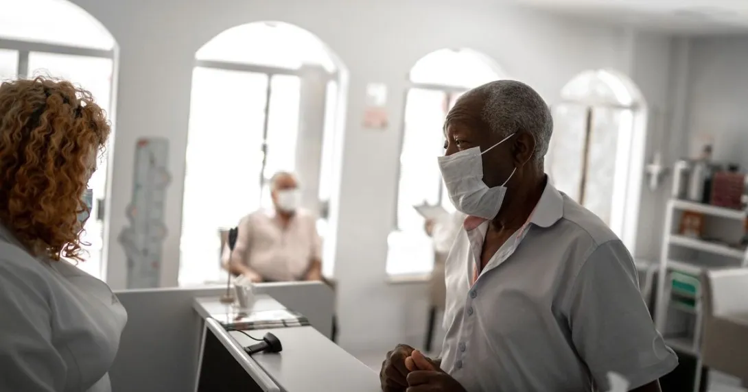 An older patient wearing a mask checking in a with a receptionist