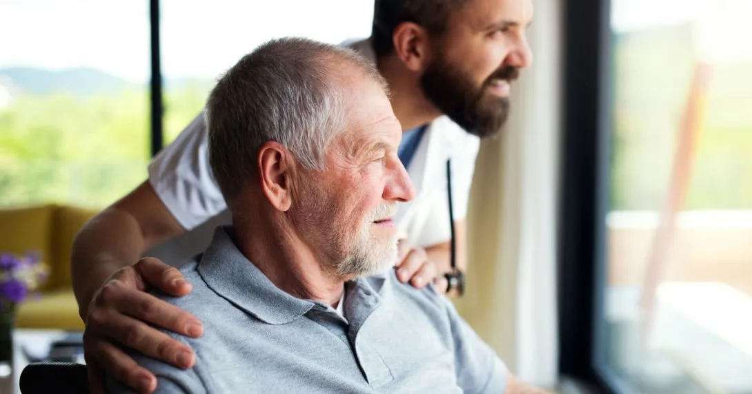 Person sitting in a wheelchair with a healthcare provider next to them, looking out a window