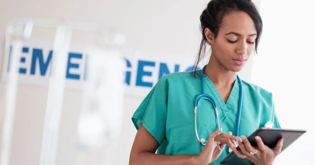 Healthcare provider wearing green scrubs and a stethoscope looking at a tablet in front of an emergency room sign