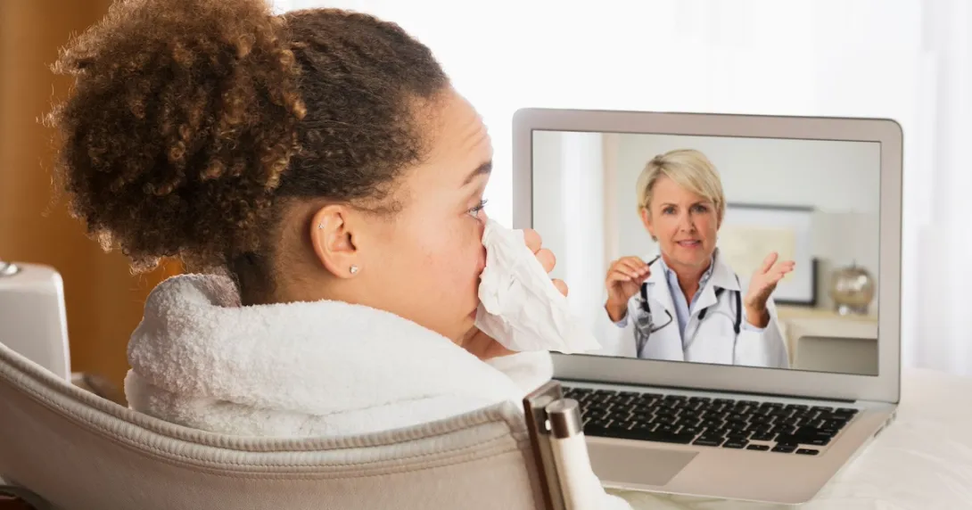 Person sitting in a chair while blowing their nose and talking to a healthcare provider on a computer