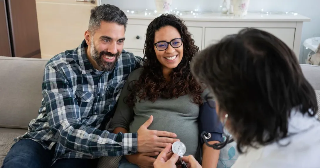 Two people sitting on a couch with one rubbing the other's stomach while both speak to a healthcare provider