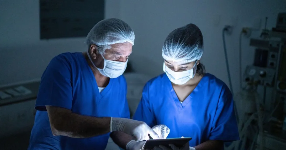 Two healthcare providers in scrubs wearing masks while in a dark room looking at a tablet