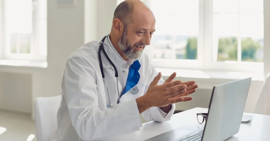 Healthcare provider sitting at a desk wearing a lab coat and a stethoscope while speaking to someone on a computer